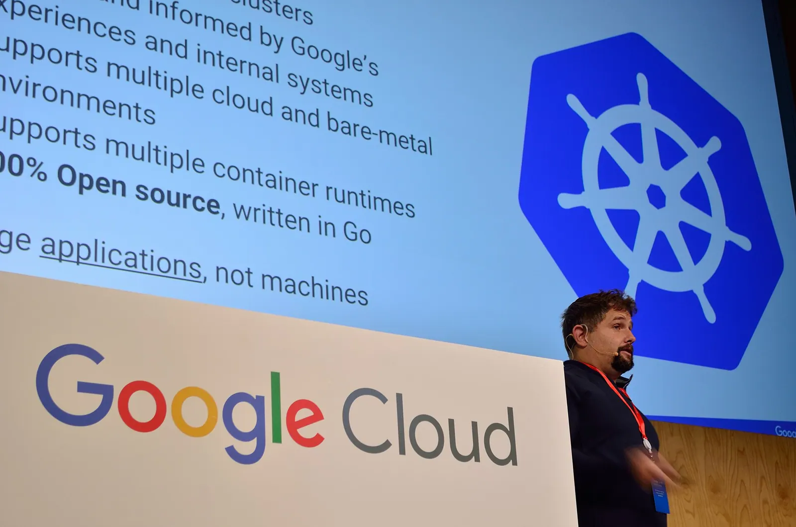Diễn giả trình bày về Kubernetes tại một hội thảo Google Cloud, phía sau là logo bánh lái Kubernetes