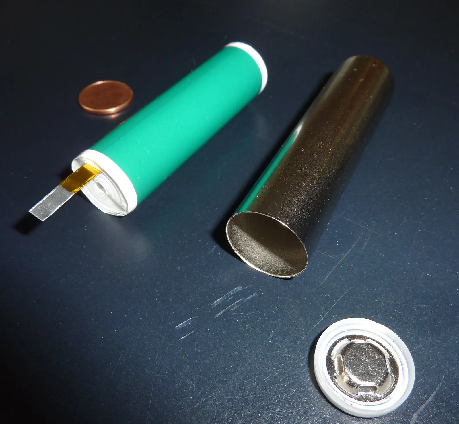 Một cell hình trụ đã tháo vỏ: lõi cuộn điện cực màu xanh nằm cạnh vỏ thép và nắp cell