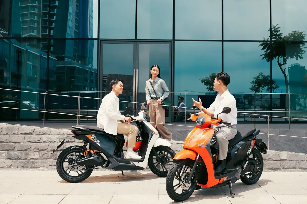 Hai người đi xe máy điện dừng trò chuyện trước toà nhà kính ở thành phố